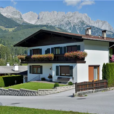 A beautiful house in the mountains with blooming plants on the balcony. In the background, majestic mountains and a clear sky stretch out.