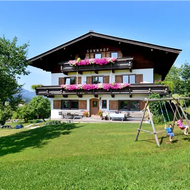 Ein hübsches Bauernhaus mit blühenden Blumenbalkonen und einer großen Wiese. Kinder spielen auf einer Schaukel im Vordergrund.