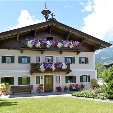 Ein traditionelles Haus mit blühenden Balkonkästen und einer schönen Gartenlandschaft. Im Hintergrund sind Berge und ein blauer Himmel sichtbar.