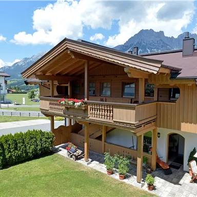 Ein gemütliches Holzhaus mit Balkon und bunten Blumen. Im Hintergrund sind die Berge und ein klarer Himmel sichtbar.