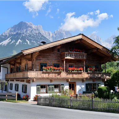 Ein traditionelles Holzhaus mit einem Balkon voller Blumen. Im Hintergrund sind majestätische Berge und ein blauer Himmel zu sehen.