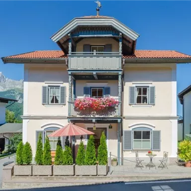 A beautiful, traditional house with blue shutters and a balcony adorned with flowers. In the foreground are tables and chairs under a sunshade.
