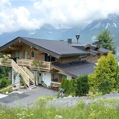 Ein charmantes Holzhaus inmitten von grünen Wiesen und Bergen. Die Umgebung ist malerisch mit einem blauen Himmel und Wolken.