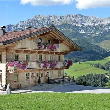 Ein wunderschönes Holzhaus mit bunten Blumenbalkonen. Im Hintergrund erstrecken sich grüne Wiesen und beeindruckende Berge unter einem klaren Himmel.