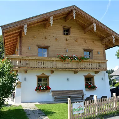Ein traditionelles, holzverkleidetes Haus mit Balkon und bunten Blumen. Der sonnige Tag verleiht der Szene eine einladende Atmosphäre.