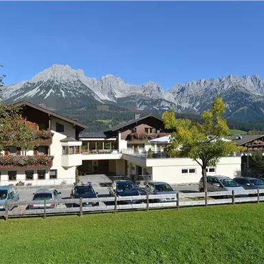 Ein modernes Gebäude mit mehreren Fahrzeugen vor einer schönen Berglandschaft. Im Hintergrund sind hohe Berge und ein klarer blauer Himmel zu sehen.