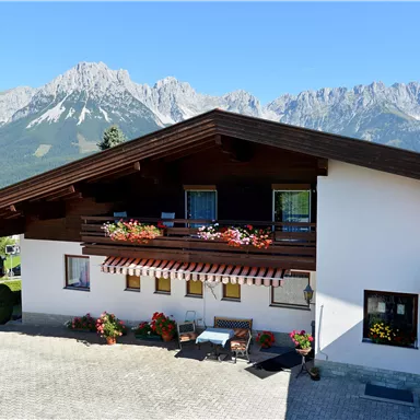 Ein charmantes Haus mit einem Holzüberstand und blühenden Blumen auf dem Balkon. Im Hintergrund sind majestätische Berge und ein klarer blauer Himmel zu sehen.