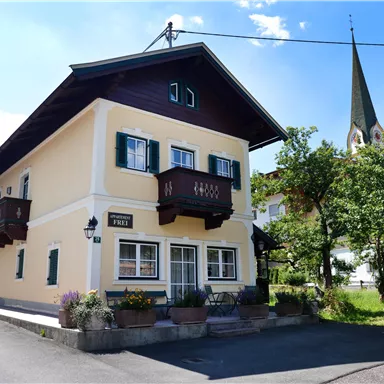 Ein charmantes Haus mit einem Balkon und grünen Fenstern. Im Hintergrund sieht man einen Kirchturm und Bäume.