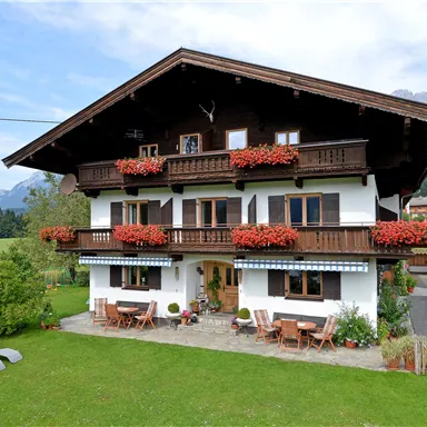 Ein charmantes alpenländisches Haus mit Holzverkleidung und Balkonen voller Blumen. Die Umgebung ist grün und bietet einen schönen Ausblick auf die Berge.