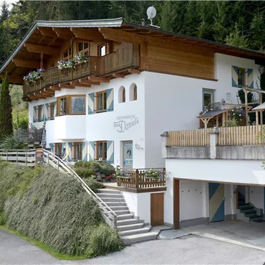 Ein modernes Haus in einer hügeligen Landschaft, umgeben von Bäumen. Das Gebäude hat einen Balkon und eine Garage.