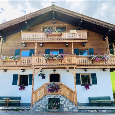 Ein typisches alpenländisches Holzhaus mit einem schönen Balkon und bunten Blumen. Im Vordergrund sind Sitzbänke zu sehen und die Umgebung ist grün.