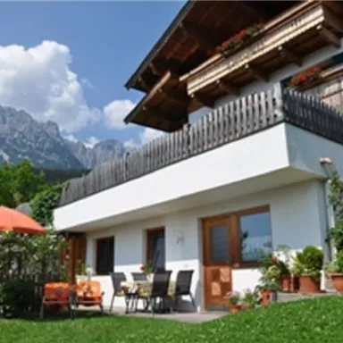 Ein charmantes Haus mit einem Balkon und einer Terrasse, umgeben von grüner Wiese und bepflanzten Töpfen. Im Hintergrund sind majestätische Berge und ein klarer blauer Himmel zu sehen.