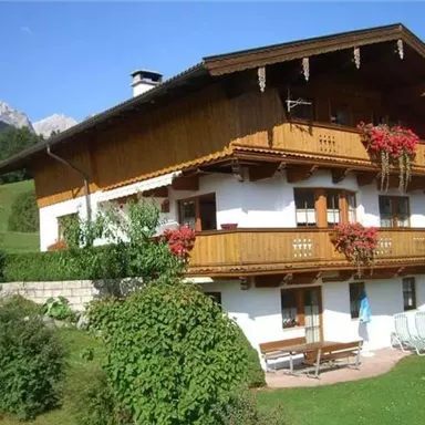 Ein gemütliches, traditionelles Holzhaus mit blühenden Balkonen. Umgeben von grünen Wiesen und Bergen im Hintergrund.