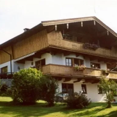 Ein traditionelles Landhaus mit Holzverkleidung und Balkonen. Es steht in einer grünen Umgebung mit Bäumen und Sträuchern.