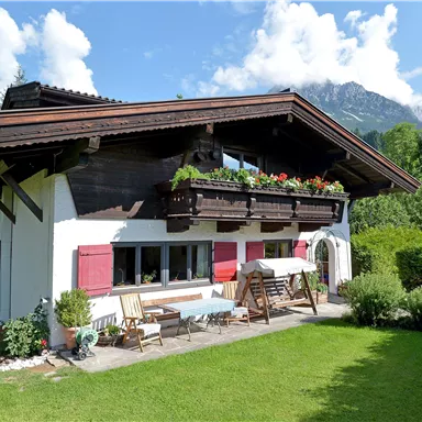 A charming country house with a well-maintained garden. In the background, green trees and mountains can be seen.