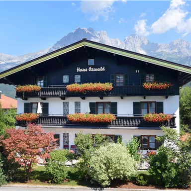 A charming building in alpine style with balconies and colorful flower boxes. In the background, majestic mountains and a clear blue sky can be seen.
