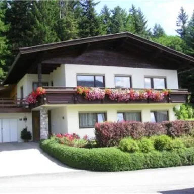 Ein schönes Haus mit einem Holzdach und bunten Blumenbalkonen. Umgeben von grünen Büschen und Wald.