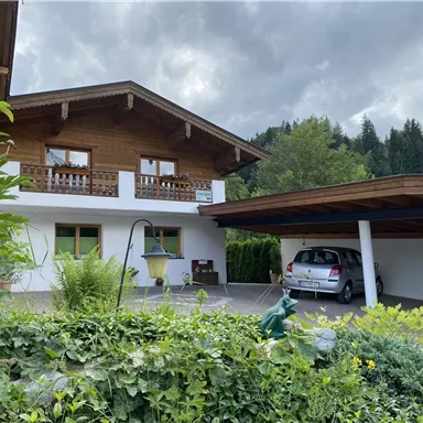 Ein traditionelles Haus im Alpenstil mit einem Holzbalcon und einer überdachten Einfahrt. Im Vordergrund sind üppige Pflanzen und ein Fahrzeug sichtbar.