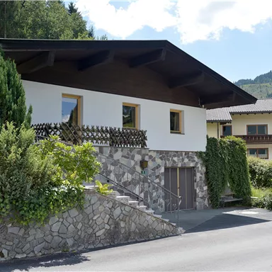 A modern house with a stone floor and plenty of greenery. In the background, additional buildings and a beautiful mountain landscape can be seen.