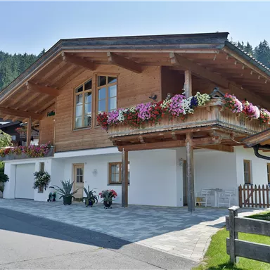 A traditional wooden house with colorful flowers on the balcony. In the background, forested hills can be seen.