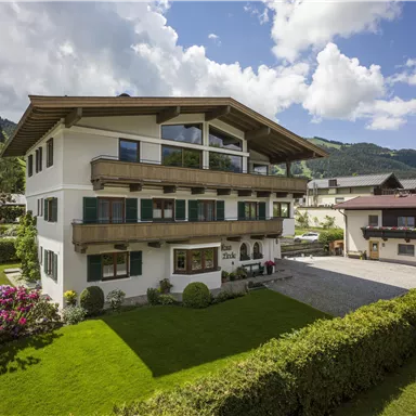 Ein modernes Gebäude umgeben von Grünflächen und Blumen. Im Hintergrund ist ein Kirchturm und die Berge zu sehen.