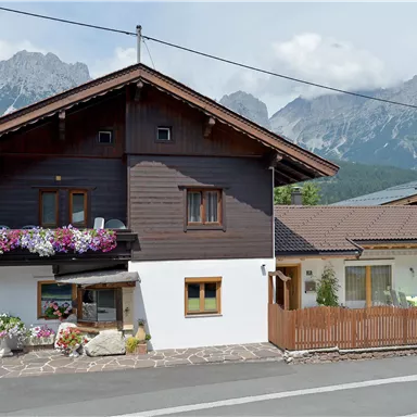 Ein traditionelles Haus mit Blumen an den Fenstern. Im Hintergrund sind beeindruckende Berge zu sehen.