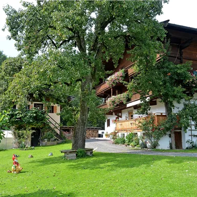 An idyllic country house with balconies and flowers on the property. Surrounded by green lawn and large trees.