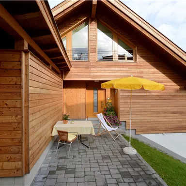 Ein modernes Holzhaus mit einem kleinen Terrassenbereich. Ein Tisch mit einem Sonnenschirm steht vor der Haustür.