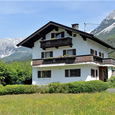Ein schönes, zweigeschossiges Haus in den Alpen, umgeben von grünen Wiesen. Im Hintergrund ragen majestätische Berge in den klaren Himmel.