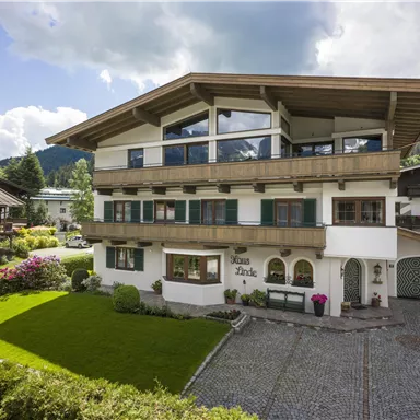 Ein modernes Haus mit einem großen Balkon und einer schönen Gartenlandschaft. Die Umgebung ist grün und von Bergen umgeben.