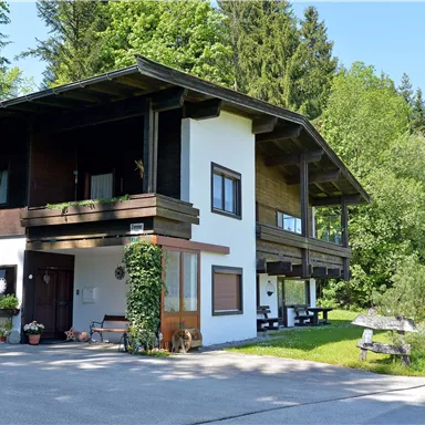 Ein charmantes Haus in ländlicher Umgebung, umgeben von Bäumen und Gras. Der klare Himmel und die Sonne sorgen für eine angenehme Atmosphäre.