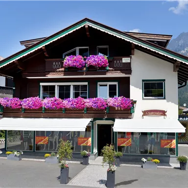 Ein schönes altes Haus mit Balkonen, dekoriert mit bunten Blumen. Im Hintergrund sind Berge und ein blauer Himmel zu sehen.