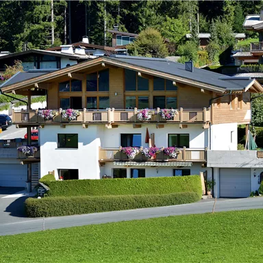 Ein modernes Haus mit mehreren Balkonen und Blumen. Die Umgebung besteht aus grünen Wiesen und weiteren Wohnhäusern.