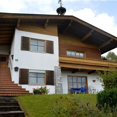 Ein charmantes Haus im alpinen Stil mit einer Holzveranda. Der Eingang ist von grünen Pflanzen umgeben und der Himmel ist leicht bewölkt.