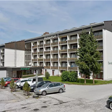 A modern hotel building with several floors and balconies. Several cars are parked in front of the hotel in a green environment.