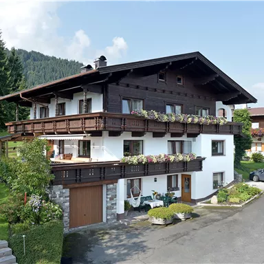 A traditional wooden house in a rural setting with many flowers on the balconies. In the background, green trees and mountains can be seen.