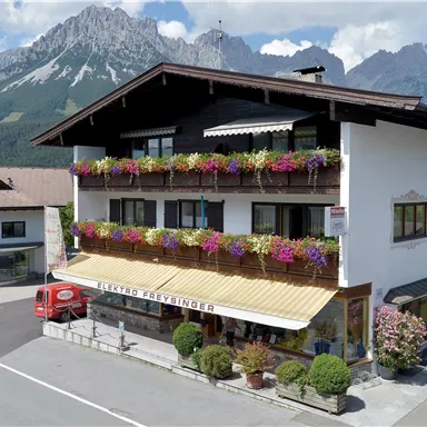 A cozy house with colorful flower balconies, surrounded by the mountains. The street runs alongside the building, which exudes an inviting atmosphere.