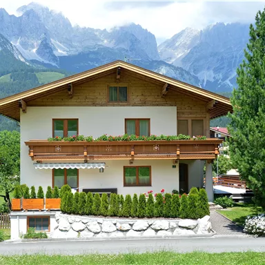A charming house with a wooden porch and blooming plants. In the background, majestic mountains and a clear sky stretch out.