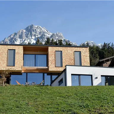 A modern house on a meadow with large windows and wooden cladding. In the background, majestic mountains and green forest can be seen.