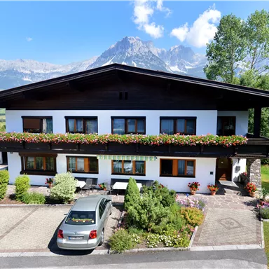 Ein charmantes Haus mit Blumenfenstern und einem Carport. Im Hintergrund sind majestätische Berge und ein blauer Himmel zu sehen.