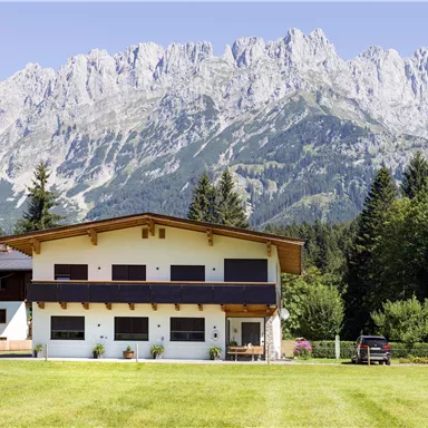 Ein modernes Haus mit einer Wiese und Bäumen im Vordergrund. Im Hintergrund erheben sich beeindruckende Berge unter einem klaren Himmel.