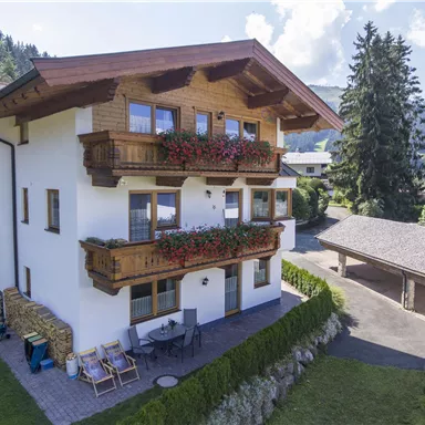 A beautiful, modern house with wooden accents and balconies, surrounded by nature. In the foreground, there is a well-maintained garden and a covered terrace.