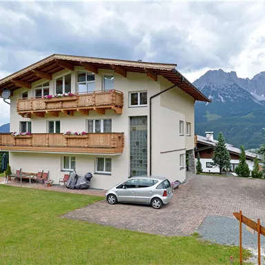 A modern house with balconies and a green lawn area. In the background, mountains are visible under a cloudy sky.