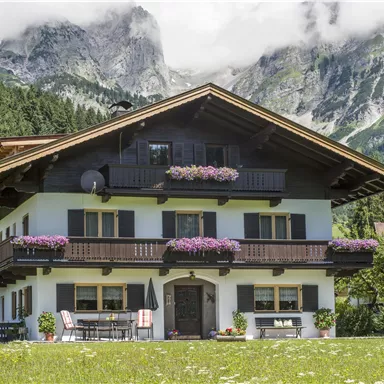 Ein traditionelles alpenländisches Haus mit Blumen auf dem Balkon. Im Hintergrund sind beeindruckende Berge und Wälder zu sehen.