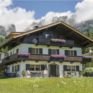 Ein traditionelles Alpenhaus mit blühenden Pflanzen und einem gepflegten Garten. Im Hintergrund sind majestätische Berge und ein blauer Himmel zu sehen.