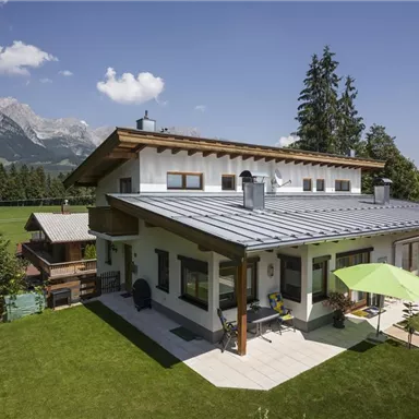 A modern house with a well-kept garden and a terrace. In the background, mountains and a clear sky can be seen.
