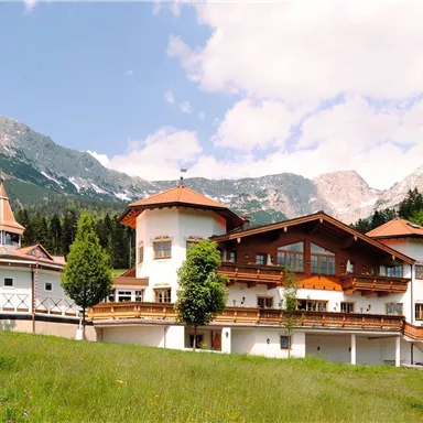 Ein malerisches Haus in den Bergen, umgeben von grünen Wiesen und Bäumen. Im Hintergrund sind beeindruckende Berggipfel und ein klarer Himmel zu sehen.