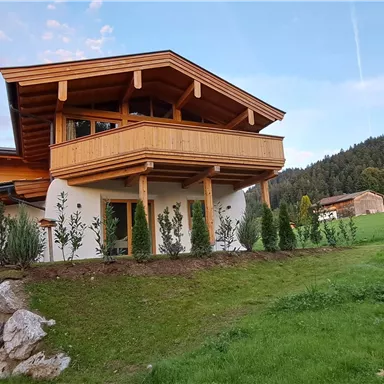 A modern house with wooden cladding in a green landscape. In the background, gentle hills and a picturesque sky can be seen.
