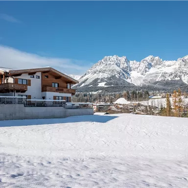Ein modernes Holzhaus in einer verschneiten Landschaft. Im Hintergrund ragen majestätische Berge unter einem klaren blauen Himmel empor.