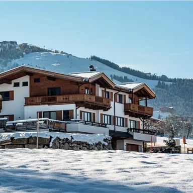 Ein schönes, schneebedecktes Haus in den Bergen. Im Hintergrund sind schneebedeckte Hügel und ein klarer blauer Himmel zu sehen.